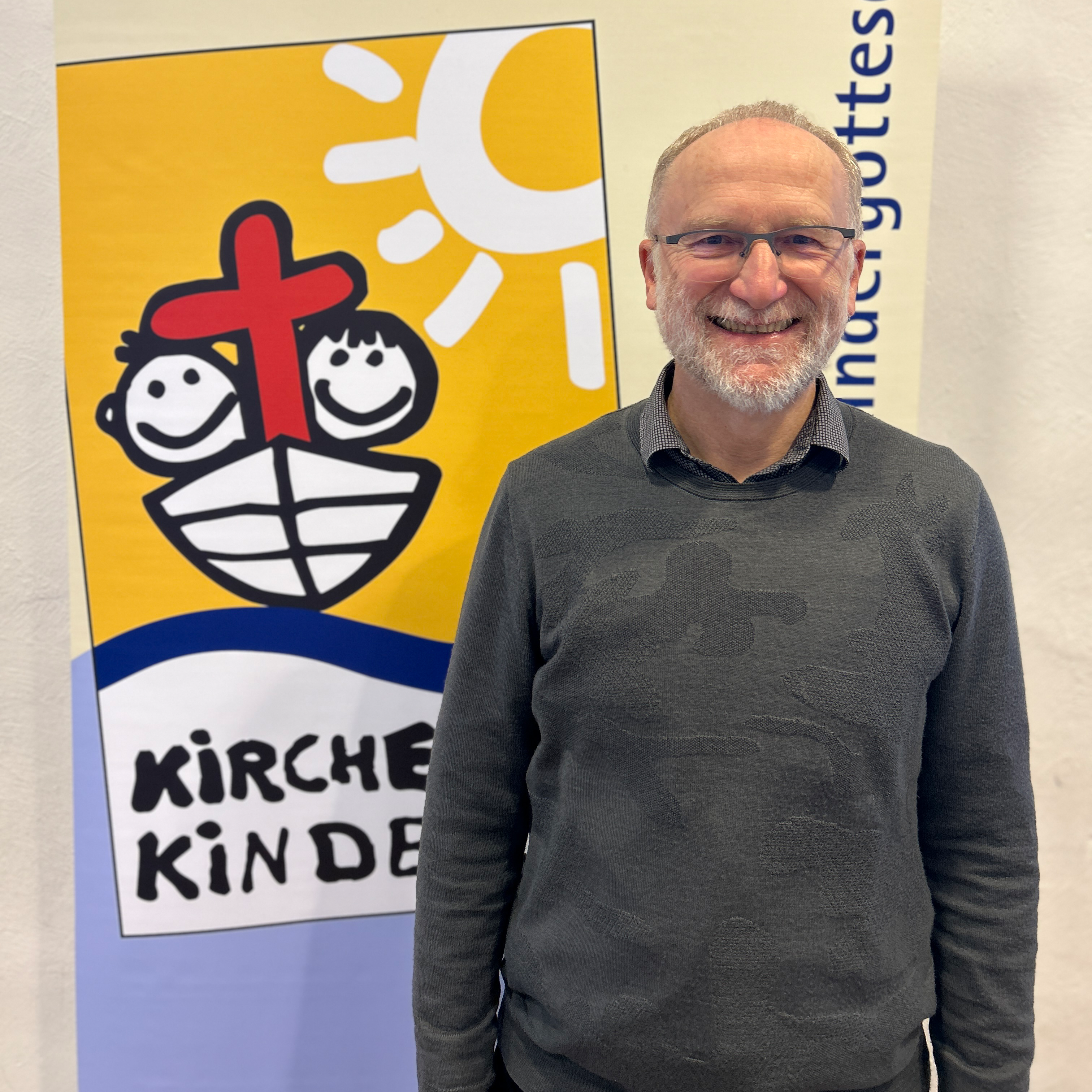 Jochen - Älterer Mann mit Halbglatze weißem Bart und Brille in einem schwarzen Pullover vor dem westfälischen Kirche mit Kindern Logo