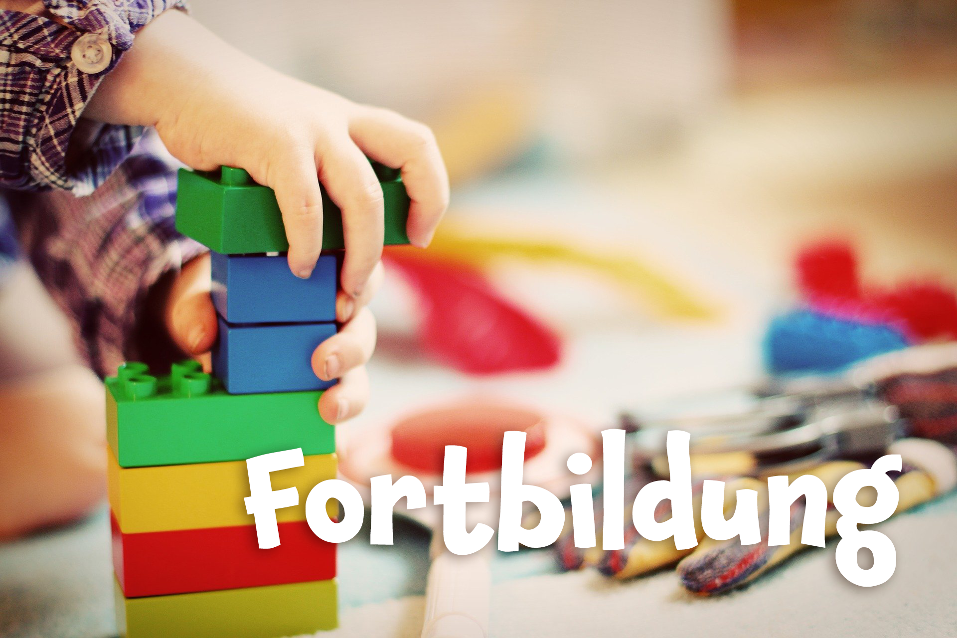Kasten mit dem Schriftzug Fortbildung. Im Hintergrund sieht man eine Kinderhand, die einen Turm aus bunten Duplo-Klötzen baut.